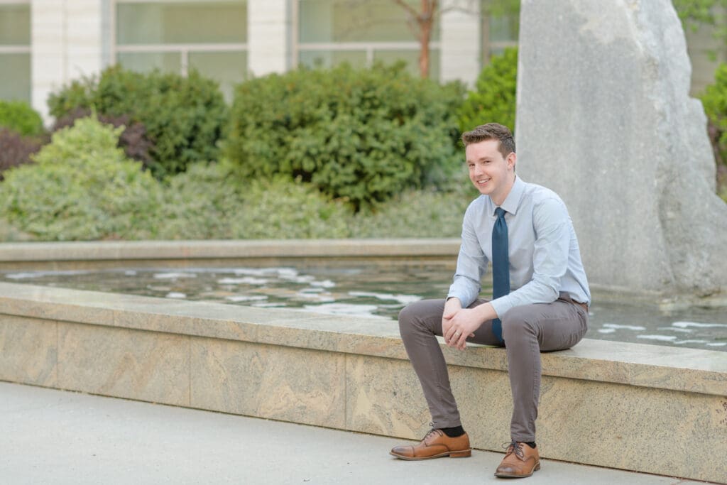 A man sits by a fountain smiling during his BYU senior portraits