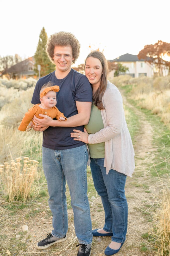 Family photo of a young father holding his infant daughter with his wife looking over his shoulder smiling during a full session with Salt Lake Photographer Jodi Cornwall Photography.