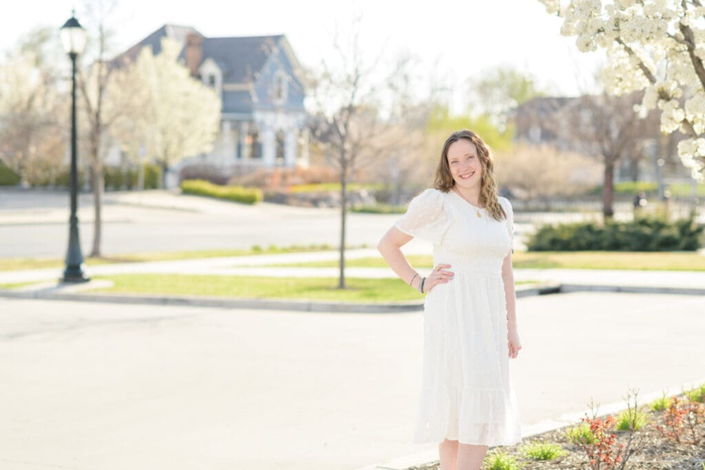 A woman in a white dress smiles as she stands by a tree in bloom during her joyful spring downtown senior session with Jodi Cornwall Photography