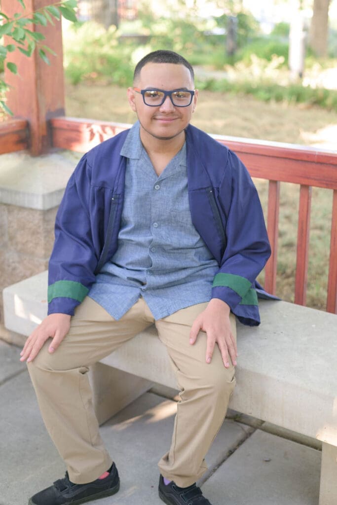 A high school graduate sits on a bench in his graduation robes during his conservation garden senior photos.